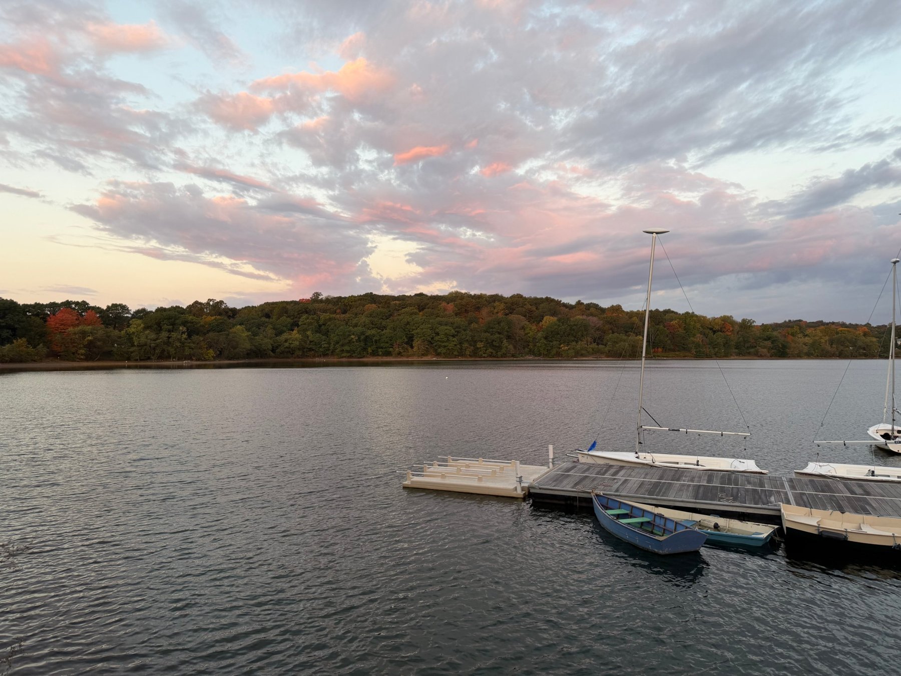 Sailboats docked on a lake at dusk, with autumn trees along the far shore